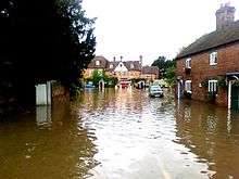Photograph showing severe flooding to a road and housing; cars are partly submerged and fire appliances (including a dinghy) are in the distance