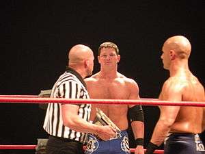 Three adult males standing in a wrestling ring. One is bald wearing a black and white striped shirt holding a championship belt. Another is staring at him wearing blue wrestling gear. The third is doing the same but is wearing black wrestling gear and is bald.