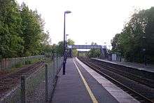 Photograph of a railway station plaform, showing the main two pairs of rails, a footbridge, and an old rusted siding