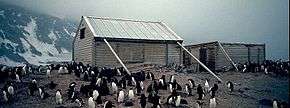  Two wooden structures surrounded by penguins. The larger, on the left, has a pitched roof and is supported by timber braces. The smaller, on the right, has no roof. Snowy slopes are visible in the background.