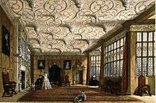 A large room with an elaborate textured ceiling. The walls are oak-panelled, and there are a number of Davenport family portraits. There is a fireplace on the left, and two large bay windows on the right. There is a chair in the closest window, and there are a few people in the room.