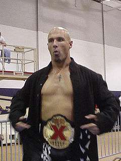 An adult white male wearing black trunks and jacket, no shirt, and a black, gold, and red championship belt at a wrestling event.