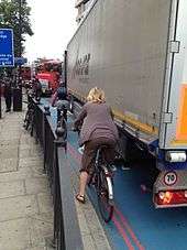 Lorry stands on blue-painted road; cyclist is between lorry and pavement with railings.