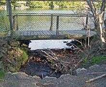 A small stream with some downed branches in it flows out of a lake at the rear, underneath a chainlink fence with a small wooden bridge behind it to a concrete culvert in the foreground