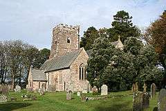 Stone building with square tower. Trees are to the right and behind with gravestones in the foreground.