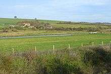 Photograph of grassland bisected by a canal, with farm buildings in the distances and the bushes of a railway cutting in the foreground.