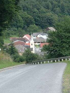 Foreground view shows two-lane paved road with low guard-rail curving downhill to the left. In the middle ground is a church with a square white bell tower and spire, and about seven other houses are visible. In the background is a forest of green conifers.