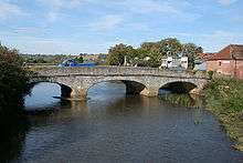 A stone three-arch bridge over water. On the bridge is a small blue lorry. Either side of the river is vegetation and to the right of the bridge houses.
