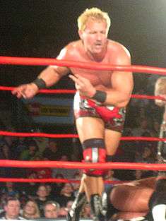 An adult male wearing red and black wrestling gear performing a strut in front of a crowd in a red-roped wrestling ring.