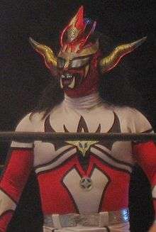 An adult male wrestler wearing a full-body costume that is red, white, and yellow. A mask covers his face, with long brown hair hanging from the back.