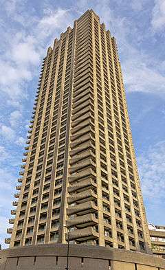 A tall concrete tower, with projecting balconies on the corners, viewed from below its lowest level. It is lit from behind and above by gold-tinges late afternoon sunlight. The sky behind it is blue with thin clouds.