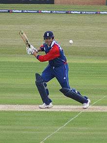 A man in a red and navy blue cricket uniform and helmet stands on a cricket pitch, watching the ball after hitting it to his left.