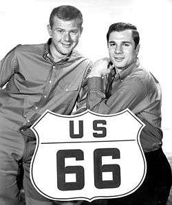 Black-and-white (greyscale) photo of blond-haired man at the left of the U.S. Route 66 large sign and black-haired man at right