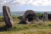 two weathered stones standing at an angle on a grassy hill, with a third doughnut-shaped stone between them