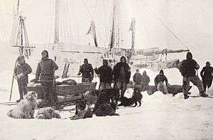 A group of men pose on the ice with dogs and sledges, with the ship's outline visible in the background