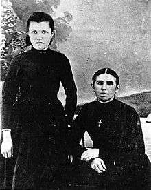 A black and white photograph of two women facing the camera. The woman on the right is seated, dressed in black and wearing a cross, with hair neatly parted in the centre. The woman on the left is standing, her hair brushed back and her left hand hidden behind her dress.