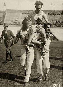 A man is carried on the shoulders of two other men. He is in a white shirt and shorts with a stylized maple leaf logo above the letters "CAN", while the two men carrying him, one black the other white, are in white long-sleeve shirts and full-length track pants