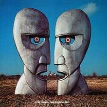 A colour photograph of two large silver-grey iron sculptures of opposing silhouetted faces. The sculptures are standing in a brown wheat field with a blue sky behind them.