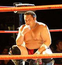 Adult Samoan male kneeling in a wrestling ring with red ropes.