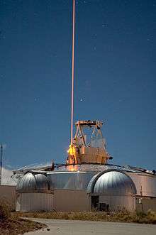 A thin bright yellow light beam goes straight up into the sky from an optical instrument