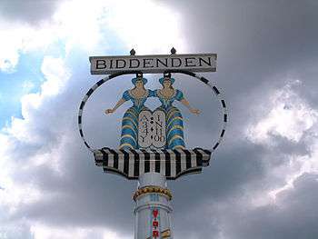 Signpost with the name of Biddenden above a circle enclosing the cut-out and brightly painted figures of two conjoined women
