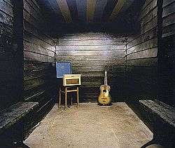 Small area with three black wooden walls; only an amplifer, speaker and guitar can be seen.