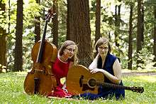 Two women sitting in woodland with a cello and a guitar.