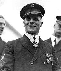 Head and shoulders of a smiling white man wearing a dark peaked cap with the letters "U.S.N" on the front and a dark jacket with three medals on the left breast and a star-shaped medal hanging from a ribbon around his neck.