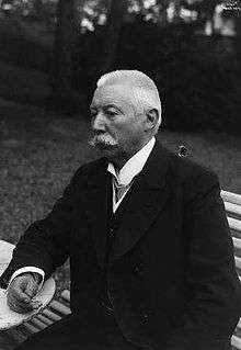 An elderly man with short white hair and a large white moustache, wearing a dark jacket, sits on a wooden bench on grass with trees in the background. He is facing to the left and looking slightly downwards.