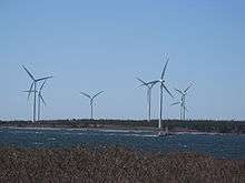 Seven three-blade wind turbines on the shore. A fishing boat is passing by. 